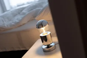 A minimalist bedroom featuring a metallic table lamp with a glowing lightbulb and a neatly made bed with white bedding.