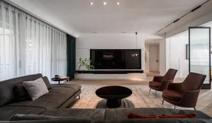 A modern living room with a gray sectional sofa, two brown armchairs, a round coffee table, and a large TV on the back wall, 