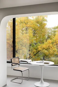 A minimalist workspace featuring a metal table and chair, with a window overlooking autumn trees.