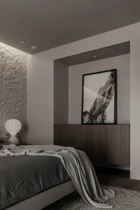 A minimalist bedroom with a textured stone wall, a bed with gray bedding, and a framed artwork.