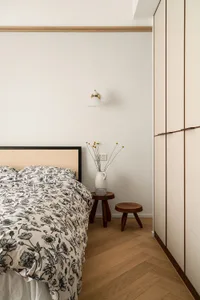 A minimalist bedroom with a wooden headboard bed, floral bedding, and a simple wooden stool with a vase of flowers.