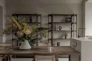 A minimalist dining room featuring a wooden table with a vase of flowers and chairs, with a bookshelf in the background.