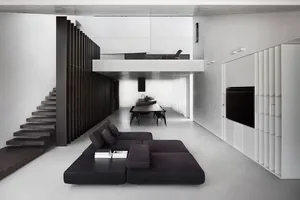 Modern minimalist living room with concrete floor, black sofa, and a raised dining area.