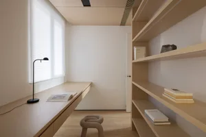A minimalist home office featuring a wooden desk and shelves with books and a black lamp.