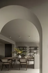 A modern minimalist dining room featuring a wooden table and chairs, with a large vase of flowers and bookshelves in the back