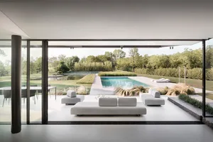 Modern residential living area with glass walls overlooking a pool surrounded by greenery.