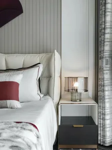 white bed in a minimalist bedroom with a black and red cushion, a glass lamp on a black drawer, a white cream and fabric wall