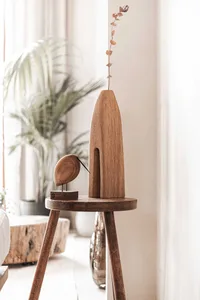A wooden stool with a vase and a bird figurine on top, set against a white wall and window with greenery.