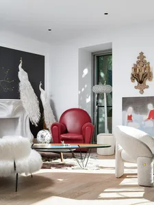 A living room with a red armchair, white fur stools, and decorative peacocks on a wooden floor with a white rug.