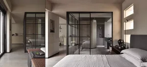 A minimalist bedroom with a concrete floor and walls, featuring a large bed and a glass-enclosed bathroom in the background.