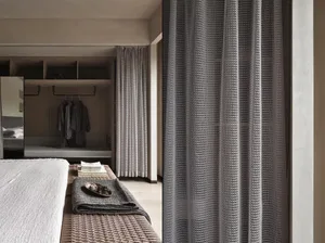 A modern bedroom with brown carpet and textured curtains, with a woven bench and a folded blanket on it.