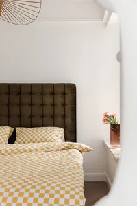 A modern bedroom featuring a velvet upholstered headboard, checkered bedding, and a vase of flowers on a windowsill.