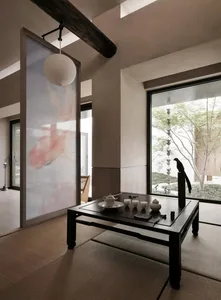 A serene living room with a minimalist tea setup on a low black table, featuring framed artwork, a lantern, and large windows