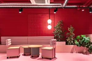 A modern cafeteria interior with pink walls and furniture, featuring industrial-style pendant lights and green plants.