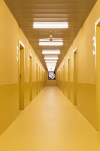 An empty, long corridor with yellow walls and doors, illuminated by overhead lighting.