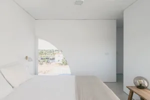 Modern minimalist bedroom with concrete walls and floor, featuring a large curved window overlooking a natural landscape.