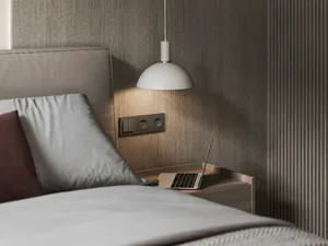 A minimalist bedroom with a neutral finish, featuring a bed and a laptop on the bedside table, illuminated by a hanging lamp.