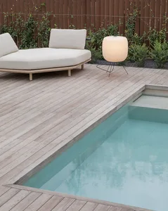 A minimalist outdoor lounge area featuring a wooden deck, glass pool, and a cushioned lounger with a round lamp.