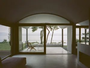 A modern interior space with glass and concrete elements, featuring a lounge chair and view of a peaceful outdoor area.