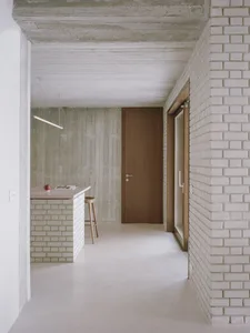 A modern minimalist kitchen with concrete walls, a wooden countertop, and a wooden stool.