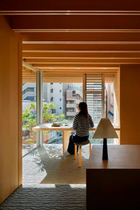 A person sitting at a wooden desk in a home office, with a chair and a lamp on a wooden table.