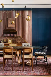 A modern, minimalist dining room featuring a wooden table and chairs, a gold chandelier, and a dark blue wall.