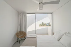 A minimalist white bedroom with a large window showing a view of a garden with palm trees.