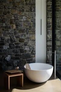 A minimalist bathroom with a stone wall, white bathtub, wooden stool, and glass door.