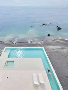 A minimalist poolside lounge area featuring glass and white tiles with a woman swimming in a clear pool.