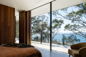 A modern bedroom featuring wood walls, a glass sliding door overlooking the sea, and a concrete floor.