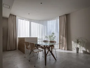 A modern dining room featuring a glass-top table, wooden chairs, and transparent curtains allowing natural light to flood the