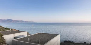 Concrete-roofed modern minimalist house situated on a cliff overlooking the sea with a sailboat in the distance.