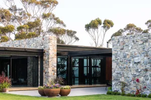 Modern residential exterior featuring stone and concrete walls, glass windows, and lush green landscaping.