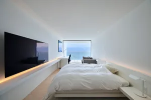 A minimalist bedroom featuring a white bed, white walls, wooden floor, and a view of the ocean through a large window.