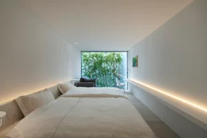 Modern, minimalist bedroom with white walls, glass window overlooking greenery, and a simple, comfortable bed.