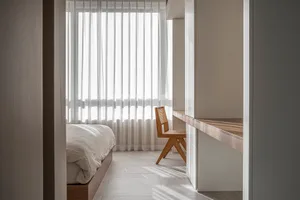 Minimalist bedroom featuring a bed with white sheets and wooden chair against a white background with vertical white curtains