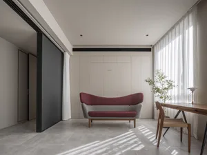 A minimalist living room with a modern sofa, wooden desk, and a plant by the window. The room features neutral-toned walls an