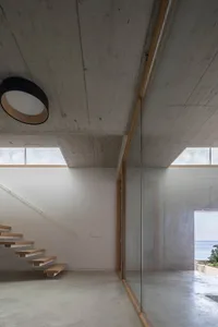 Modern minimalist interior with concrete walls and floor, featuring a wooden staircase and large windows overlooking the sea.