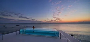 A modern poolside view with a glass infinity pool overlooking the ocean during sunset.