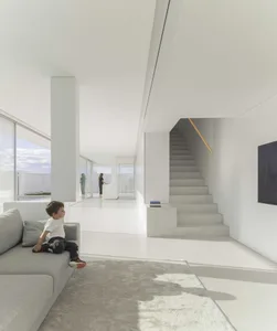 A minimalist living room with white walls, concrete floors, and a large window allowing natural light to fill the space.
