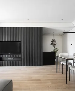 A modern living room with black wooden paneling, a flat-screen TV, and a wooden dining table with white chairs.