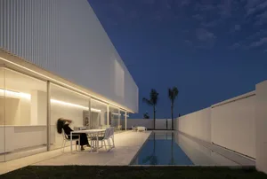 Modern house exterior at night with glass walls and concrete patio.