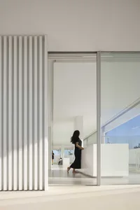 A minimalist interior space featuring white walls, glass partitions, and a woman standing in a black dress.