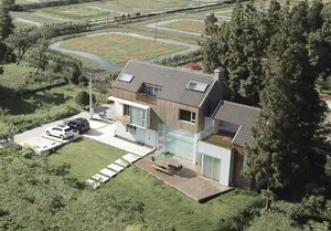 Modern residential villa with wooden frame exterior, surrounded by greenery and a pathway leading to the front door.