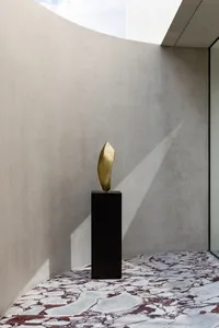 A golden abstract metal sculpture displayed on a black pedestal in a minimalist room with curved walls and marble floor.