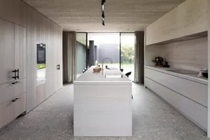 Modern, minimalist kitchen with concrete countertops and floor, featuring an island with a sink and a view of the outdoors th