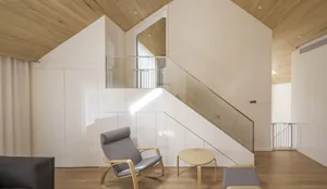 A modern minimalist living room with white walls and wood flooring, featuring a single wooden chair and table with a glass ba