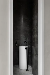 極簡浴室內，一座流線圓柱形白色洗手台，置於深色質感混凝土牆面與地面之前。