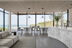 Modern residential interior with large glass windows overlooking a scenic coastal landscape, featuring a concrete and glass d