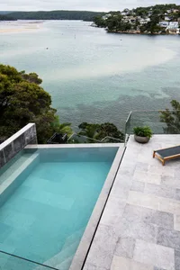 A modern poolside patio with a concrete pool and a lounge chair overlooking a serene waterfront.